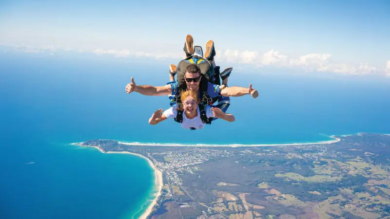 Tandem skydivers above Byron Bay coast, smiling and giving thumbs up during 15,000ft skydive with Brisbane transfer included.