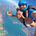 Skydivers tandem jump over Newcastle coast from 15,000 feet, smiling and flashing peace signs in exhilarating skydiving adventure.