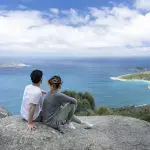 A couple relaxes atop a rocky cliff at Wilsons Promontory Day Tour, overlooking scenic sea views, islands, and lush coastline.