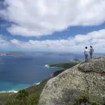 Visitors stand atop a rocky cliff at Wilsons Promontory, enjoying panoramic ocean views, scenic islands, and a dramatic cloudy sky.