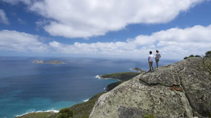 Visitors stand atop a rocky cliff at Wilsons Promontory, enjoying panoramic ocean views, scenic islands, and a dramatic cloudy sky.