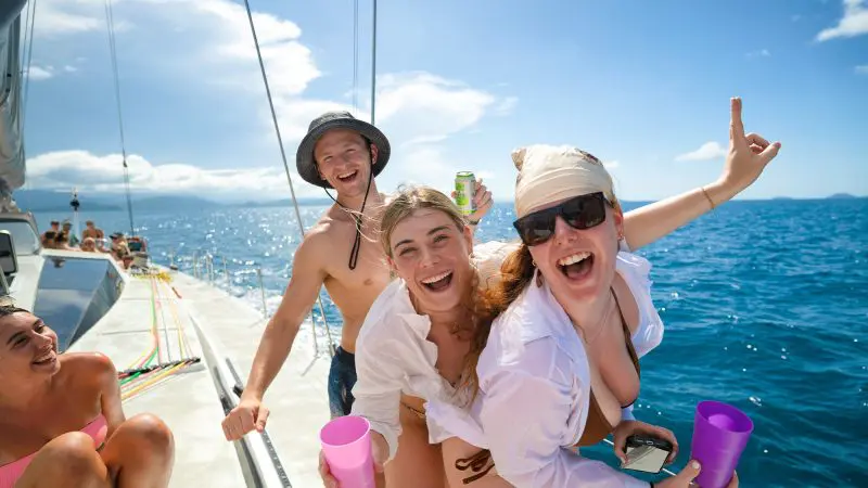 Three friends smiling on a sailboat during their 28 Day Oz East Coast Adventure, enjoying drinks and stunning ocean views in the background.
