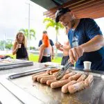 Man barbecuing sausages outdoors at 28 Day Oz East Coast Adventure Intro Travel event, highlighting group travel experience.