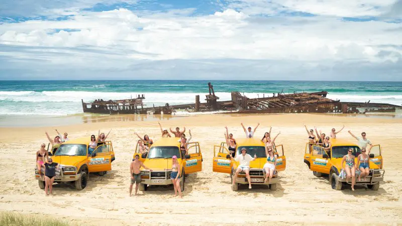 Travellers on the 28 Day Oz East Coast Adventure smile and wave beside vibrant yellow lorries at a famous rusted shipwreck in Australia.