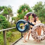 Three women in swimsuits with colourful rubber rings smile on a scenic wooden bridge during the 28 Day Oz East Coast Adventure tour.