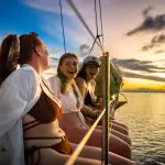 Three women in swimming costumes laughing on a sailing boat at sunset during the 28 Day Oz East Coast Adventure tour, enjoying the sea views.