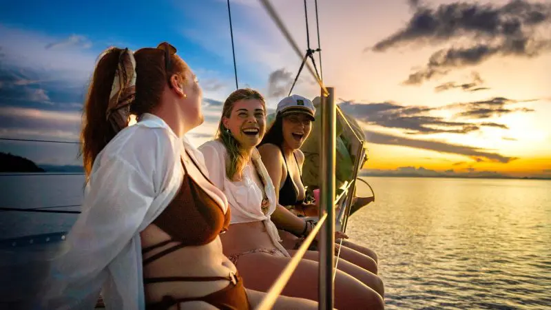 Three women in swimming costumes laughing on a sailing boat at sunset during the 28 Day Oz East Coast Adventure tour, enjoying the sea views.