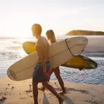 Two travellers with surfboards stroll a golden sandy beach at sunset, kicking off their 10 Day Oz Intro Sydney to Brisbane adventure tour.
