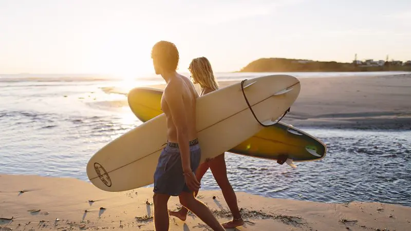 Two travellers with surfboards stroll a golden sandy beach at sunset, kicking off their 10 Day Oz Intro Sydney to Brisbane adventure tour.
