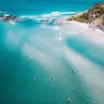 Stunning aerial shot of turquoise sea, kayakers, golden sandy beach, and rugged rocky shore on 10 Day Oz Intro Sydney to Brisbane tour.