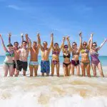 Group enjoying 10 Day Oz Intro Sydney to Brisbane tour, standing in sea waves, smiling and posing under clear blue sky for photos.