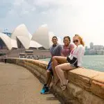 Three friends enjoy waterfront views on a stone wall during their 10 Day Oz Intro Sydney to Brisbane adventure travel experience.