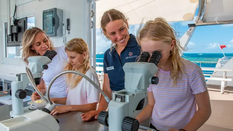 Four people, including two children, examine samples in a sunlit laboratory with panoramic views of the Reef Magic Outer Barrier Reef Pontoon.
