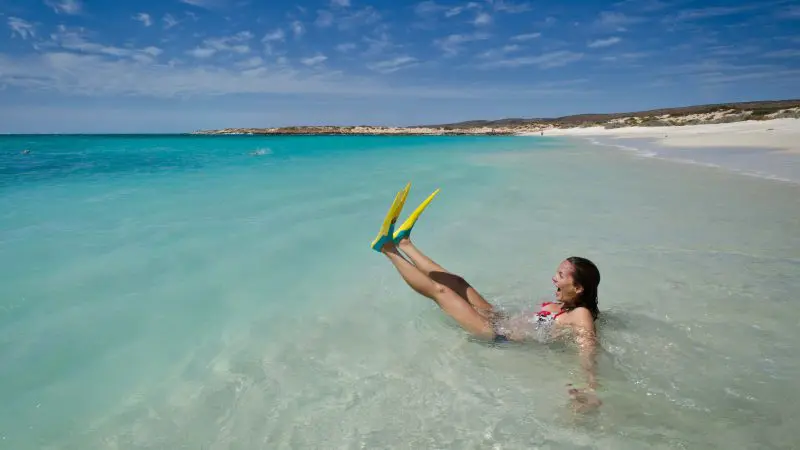 Woman wearing snorkel gear and bright yellow fins relaxes on a beach, ideal for Perth to Exmouth Return Tour Hostel Twin adventure.