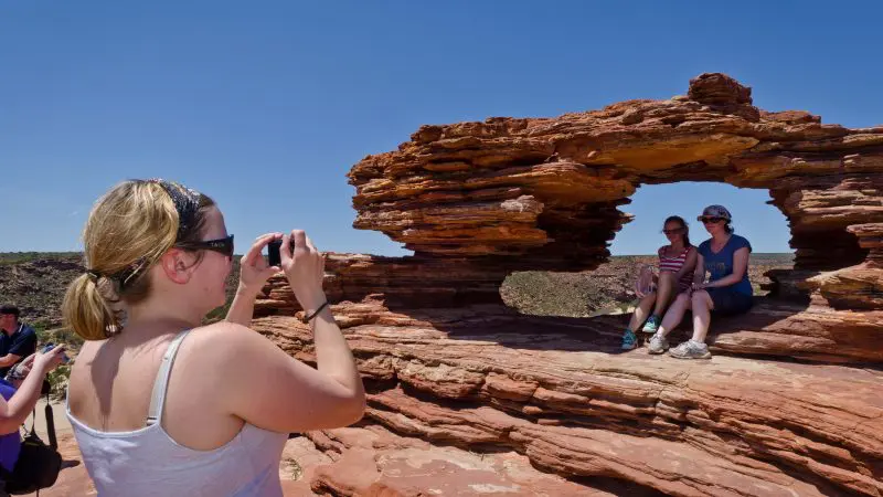 A woman takes photos of two people atop a striking rock formation with a natural arch, ideal for Perth to Darwin tours without Broome.