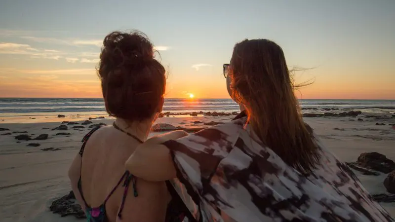 Two women relax on a rocky coast at sunset, savouring their Perth to Darwin adventure tour with Broome accommodation deals included.