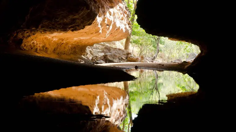 Discover a sunlit cave entrance with rugged rocky walls, lush trees, and clear water—Perth to Darwin tour with Broome stay.