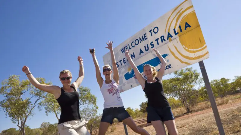 Three happy travellers jump and smile by the iconic Welcome to Western Australia sign on their epic 10 Day Broome to Darwin 4WD Kimberley tour.