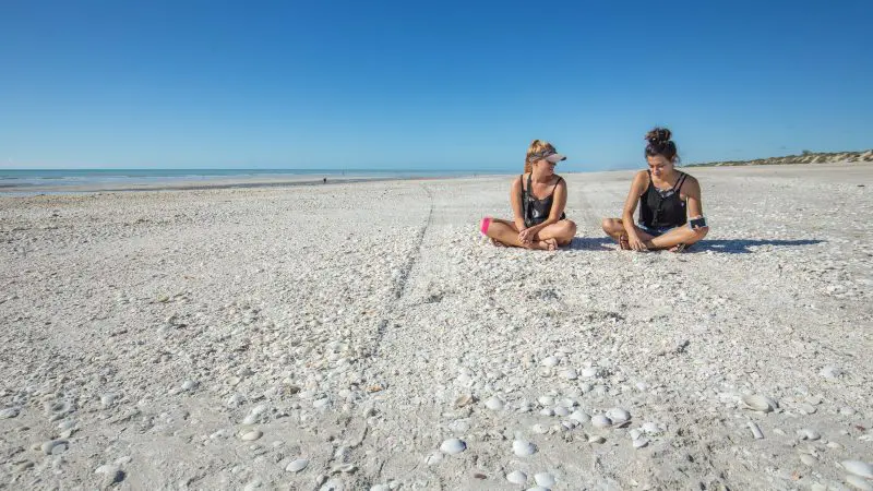 Two women smiling and chatting on a picturesque, shell-covered beach during the Darwin to Perth Adventure Tour with Broome accommodation.