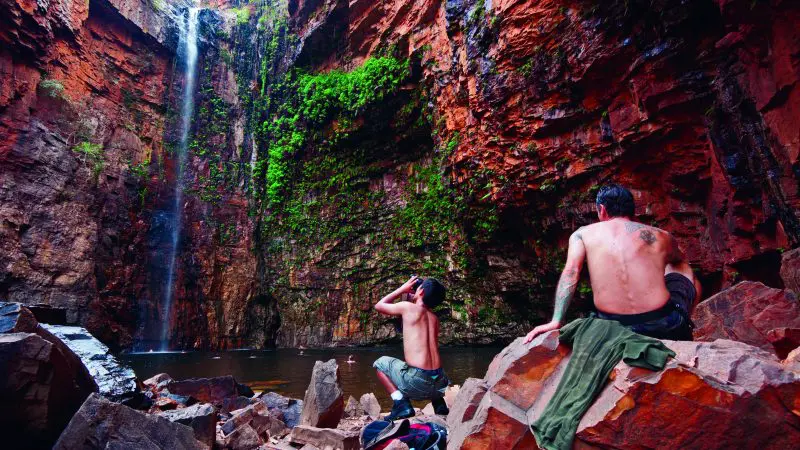 Couple relax beside a scenic waterfall in a striking red rock canyon on a Darwin to Perth Adventure Package, Broome included.