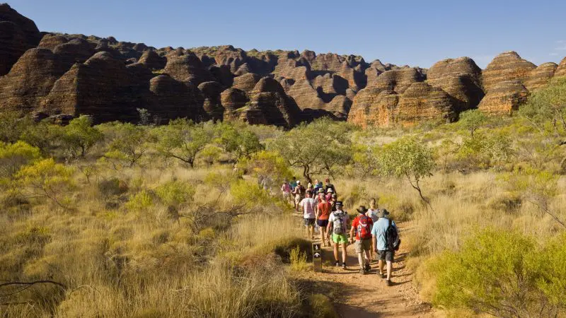 Hikers on a Perth to Darwin tour trek scenic trails through rugged rocky hills and golden dry grasslands, excluding Broome accommodation.
