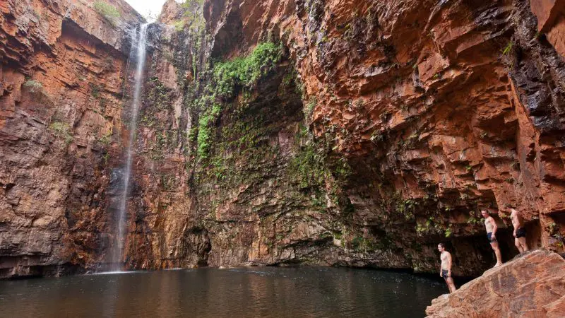 Spectacular waterfall cascades into a rocky pool as Darwin to Broome 4WD Adventure travellers enjoy panoramic views from red cliffs above.
