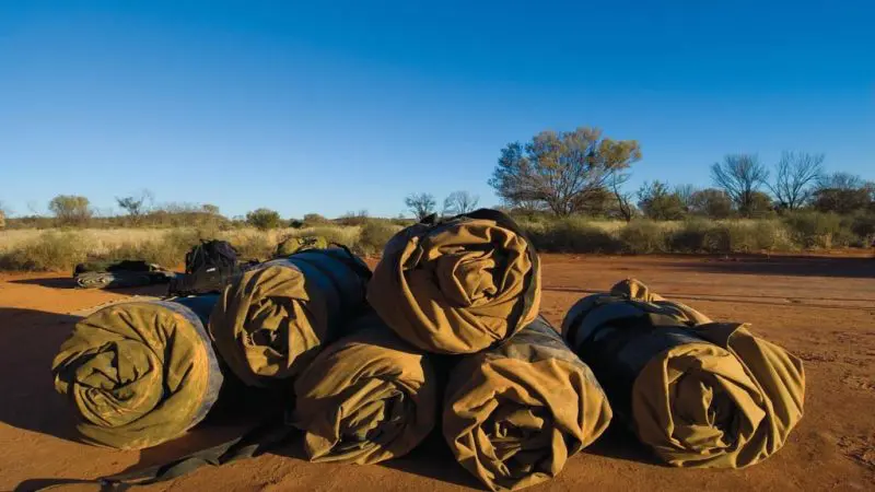 Six rolled-up swags on red outback dirt, prepared for your epic 5-day camping tour from Yulara to Alice Springs adventure.