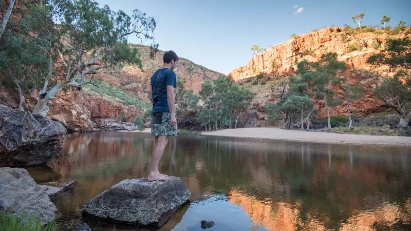 Traveller stands atop a rugged rock beside a tranquil river during the renowned 5 Day Outback Camping Adventure in Alice Springs.