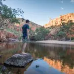 Traveller stands atop a rocky outcrop beside a tranquil river on a 5-Day Outback Camping Adventure from Yulara to Alice Springs.