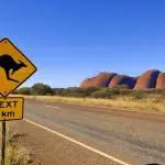 Bright yellow kangaroo road sign with "NEXT 5km" on scenic Uluru tour, Yulara loop; iconic red rock formations in the background.