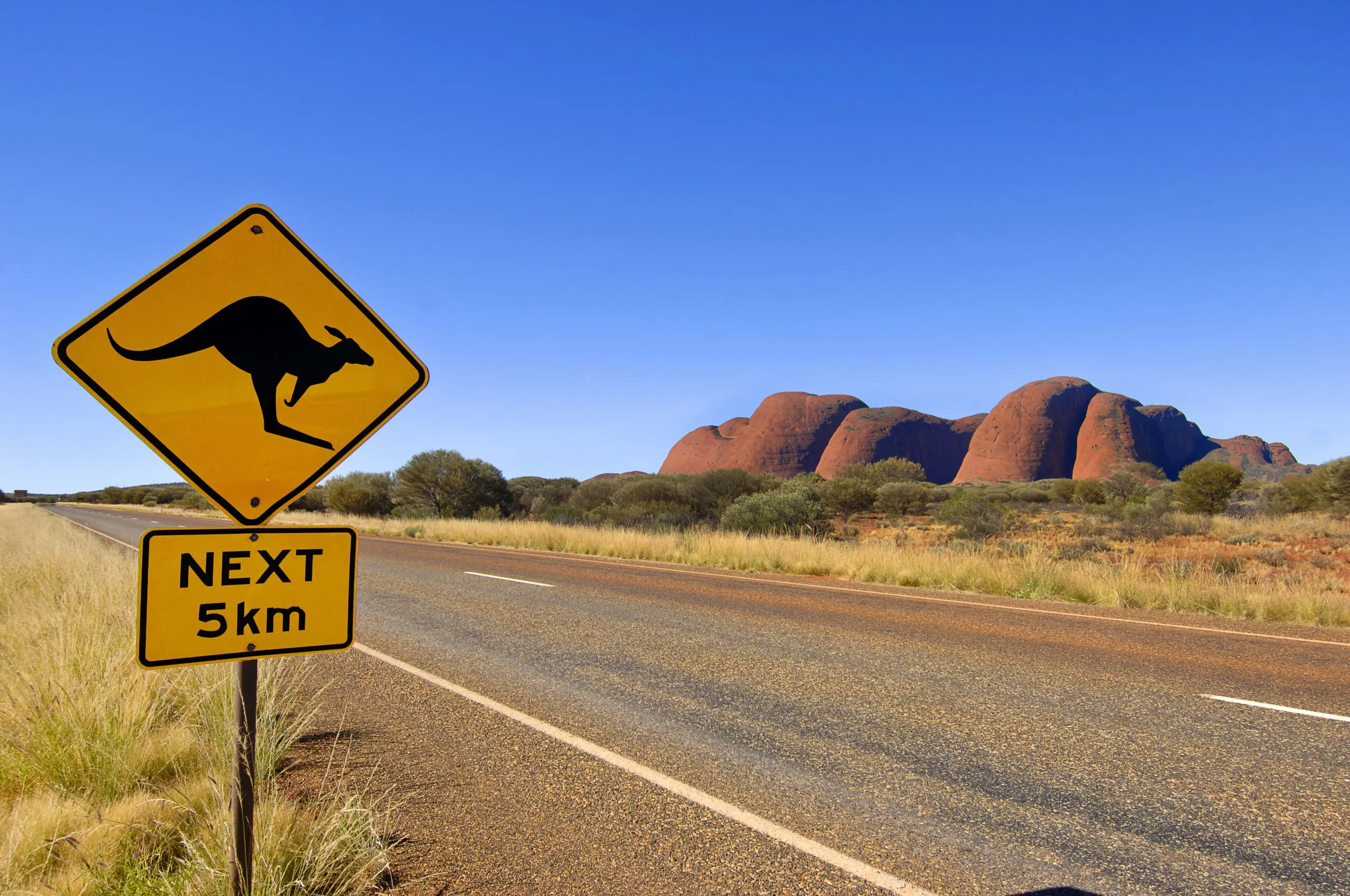 Bright yellow kangaroo road sign with "NEXT 5km" on scenic Uluru tour, Yulara loop; iconic red rock formations in the background.