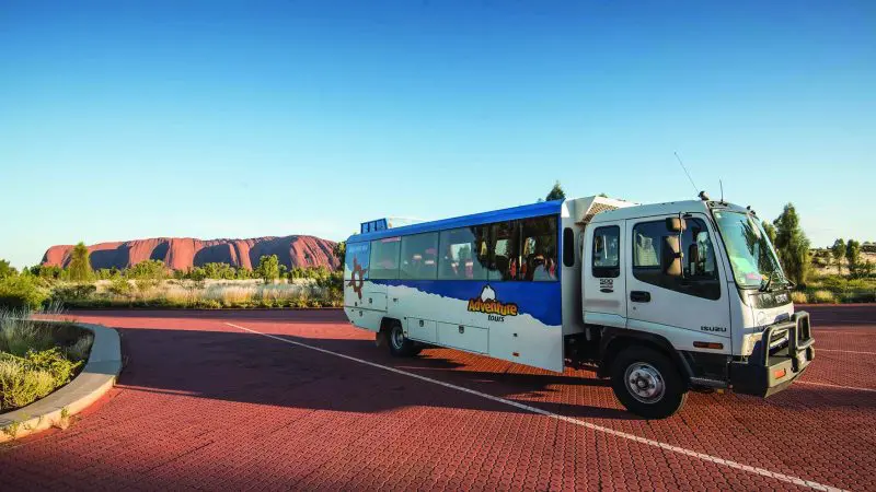 Embark on a 3-Night Uluru Adventure: coach on red-brick road near Alice Springs, Yulara, and iconic Uluru in the background.