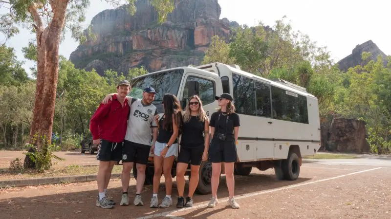 Five smiling adventurers stand in front of a large white tour vehicle on their 4 Day Kakadu, Katherine, and Litchfield Expedition, surrounded by dramatic cliffs and lush trees.
