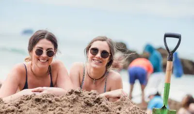 Two women in stylish outfits pose playfully on sandy ground, striking quirky Funky Chicken-inspired stances for a vibrant photoshoot.