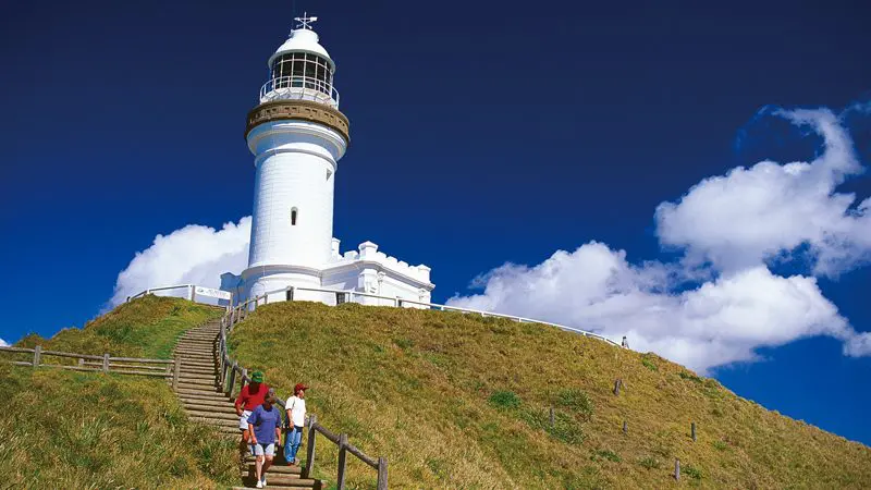 Three people relax at Byron Bay, descending steps near the iconic white lighthouse beneath a vibrant, cloud-dotted blue sky.