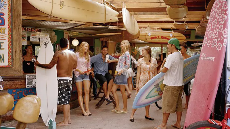 Smiling surfers with colourful boards gather and laugh by a rustic surf shop in Byron Bay, Australia’s top destination for surfing enthusiasts.