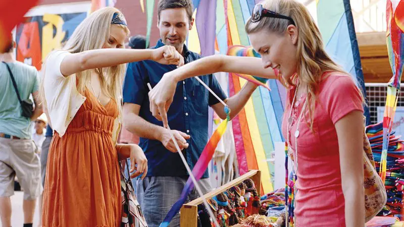 Three friends browsing vibrant handmade crafts and smiling at the lively Chill Out At Byron Bay outdoor market festival.