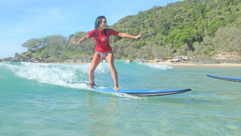 Woman in red shirt expertly surfing Longest Wave Beach, Noosa, surrounded by trees during top-rated Australia surf lesson experience.