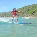 A woman in a vibrant red shirt rides Australia’s Longest Wave by a scenic sandy beach dotted with trees and parked cars in the background.