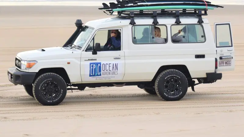 White 4WD with rooftop surfboards cruises Noosa’s Longest Wave Beach; “Epic Ocean Adventures” branding visible on the door.
