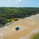 Blue van parked on a vast sandy beach at Kgari 4 Day Experience, framed by lush green forest under clear skies—ultimate adventure scene.