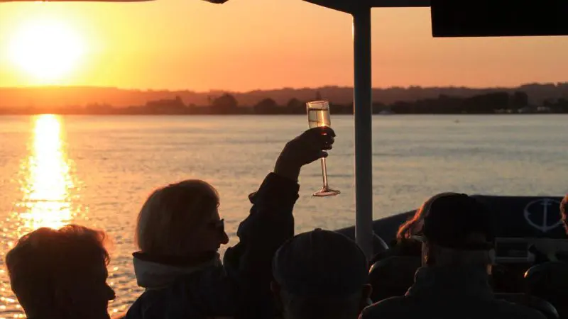 Group of people enjoying a scenic river cruise at sunset, raising glasses on a boat with glistening water and vibrant sun backdrop.