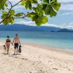 Couple in swimwear walks hand-in-hand on pristine sandy beach with Frankland Island Express Tour kit, sea and mountains in background.