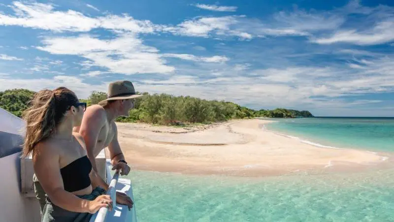 Two travellers on a boat admire pristine blue waters, white sandy beach, and clear sky during a 1 Day Frankland Islands Reef Cruise.