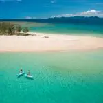 Two adventurers paddleboard in crystal-clear turquoise waters by a pristine sandy island on a Frankland Island Express Tour.