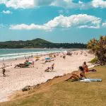 Travellers unwind on a sunlit grassy hill and white sandy beach beside the ocean, along the Sydney to Cairns East Coast adventure tour.
