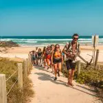 Travellers on the Fully Guided Ultimate East Coast Sydney to Cairns 5 Weeks tour hike a scenic sandy path from the beach under blue skies.