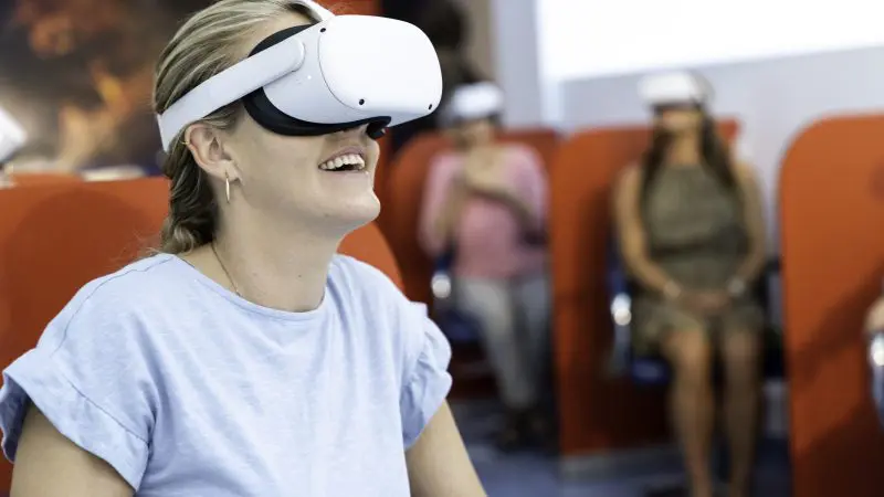 A smiling woman explores Croc N History Explorer using a VR headset, whilst others immerse themselves in virtual reality in a well-lit room.