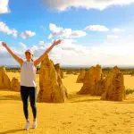 Traveller with arms raised stands amid limestone Pinnacles on a Perth day tour, dramatic clouds overhead, experiencing WA’s unique landscape.