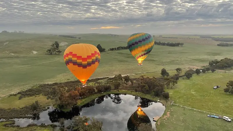 Balloon Flight With Breakfast Perth: Vibrant hot air balloons soar over tranquil Avon Valley fields, pond, and dramatic cloudy skies.
